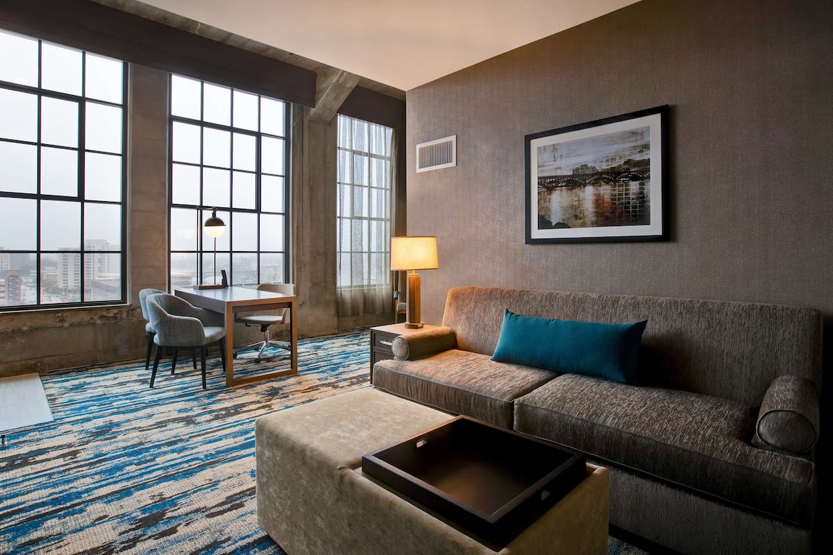 Una habitación de hotel moderna con un ventanal grande, un sofá, un escritorio y una lámpara.