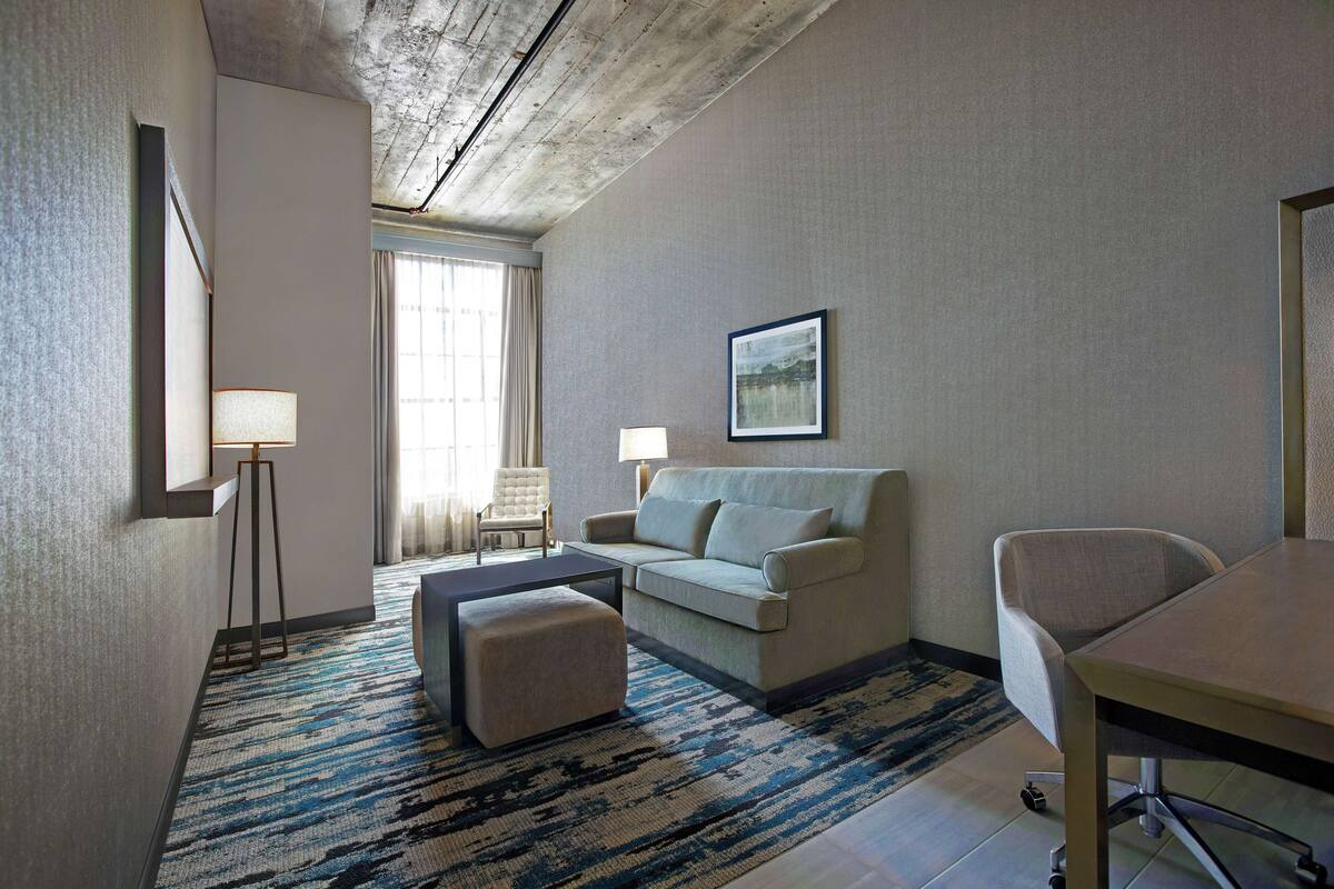 Habitación de hotel con sofá, reposapiés y una mesa de comedor. Hay una lámpara, una silla y un cuadro en la pared.