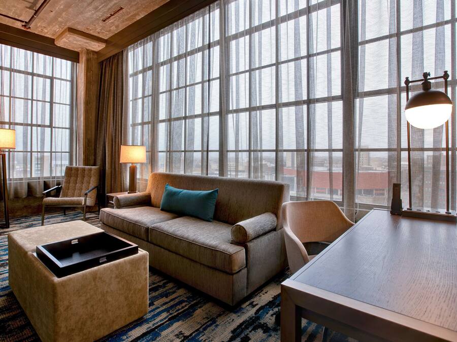 A modern hotel room with a large window, a sofa, a chair, a small table, and a lamp.