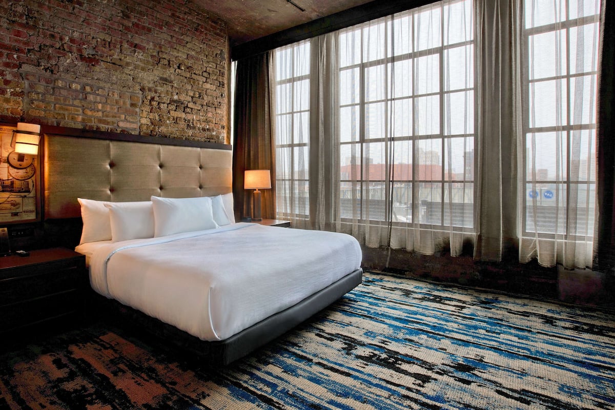 Habitación de hotel con una cama grande, pared de ladrillo y un amplio ventanal con cortinas.