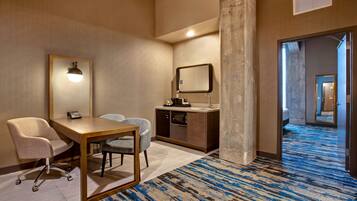Une chambre d’hôtel équipée d’un bureau, de deux chaises, d’une petite kitchenette, d’un miroir et d’une moquette à motifs.