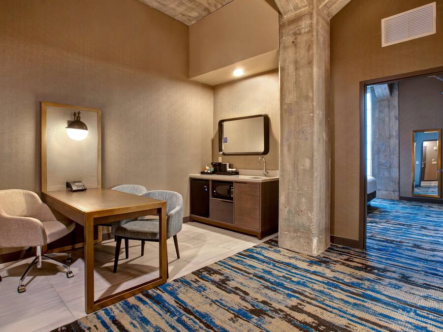 Une chambre d’hôtel équipée d’un bureau, de deux chaises, d’une petite kitchenette, d’un miroir et d’une moquette à motifs.