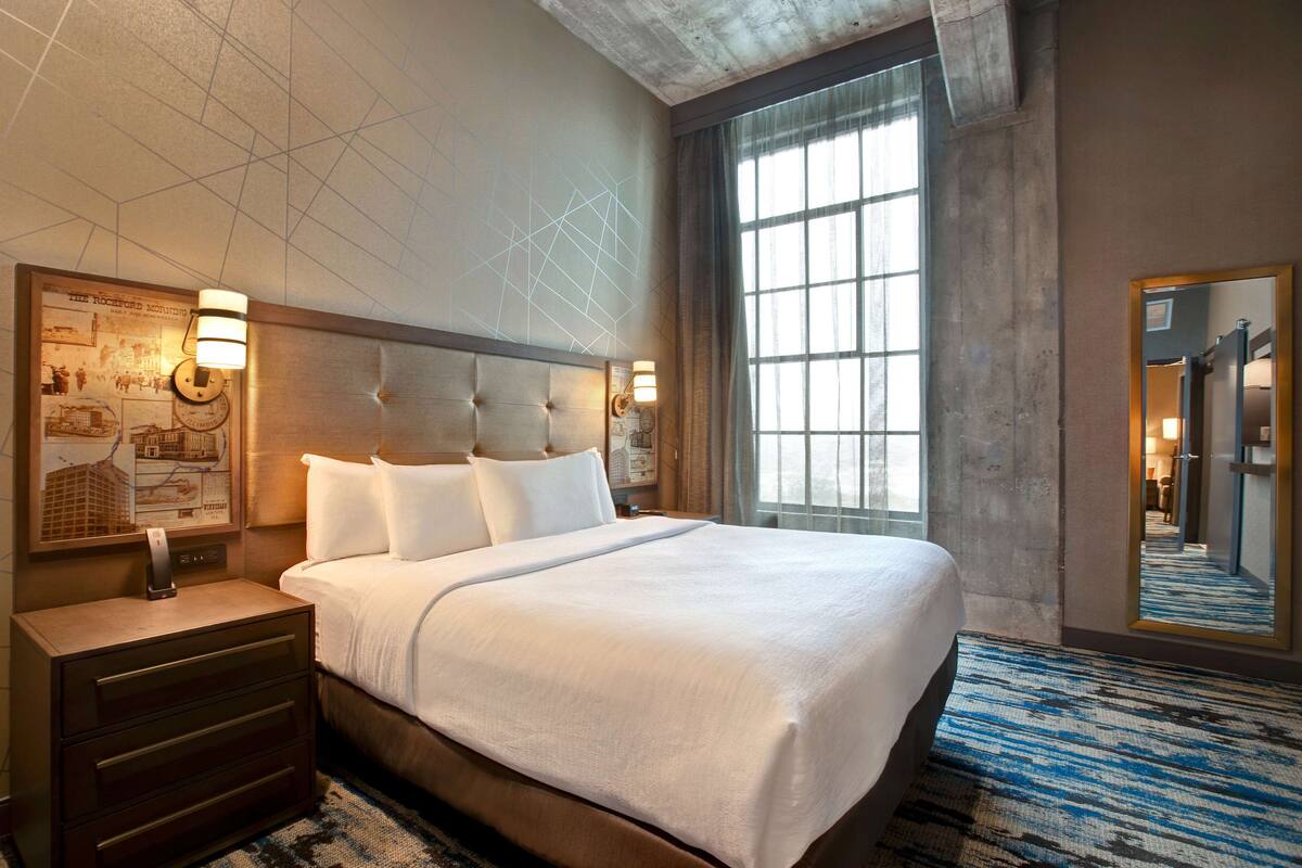 Habitación de hotel con una cama grande, una mesita de noche, una lámpara de pared y un amplio ventanal con cortinas.
