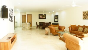 Sala de estar com sofá marrom, mesa de centro de vidro e TV fixada na parede. Ao fundo, área de jantar com mesa e cadeiras.