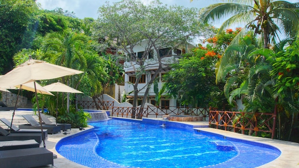 Piscina com bordas azulejadas, rodeada por espreguiçadeiras e guarda-sóis, com um edifício e árvores ao fundo.