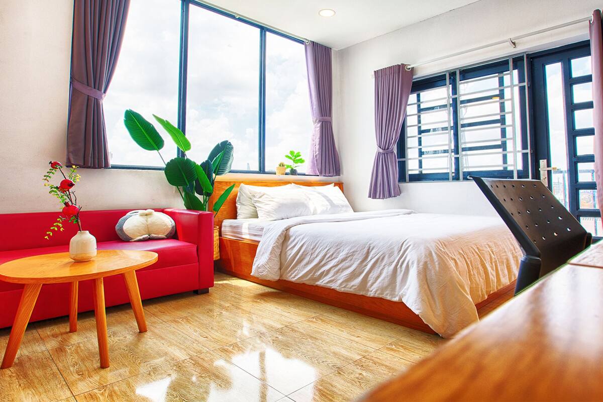 A hotel room with a bed, a red sofa, a small table, and a large window with curtains.