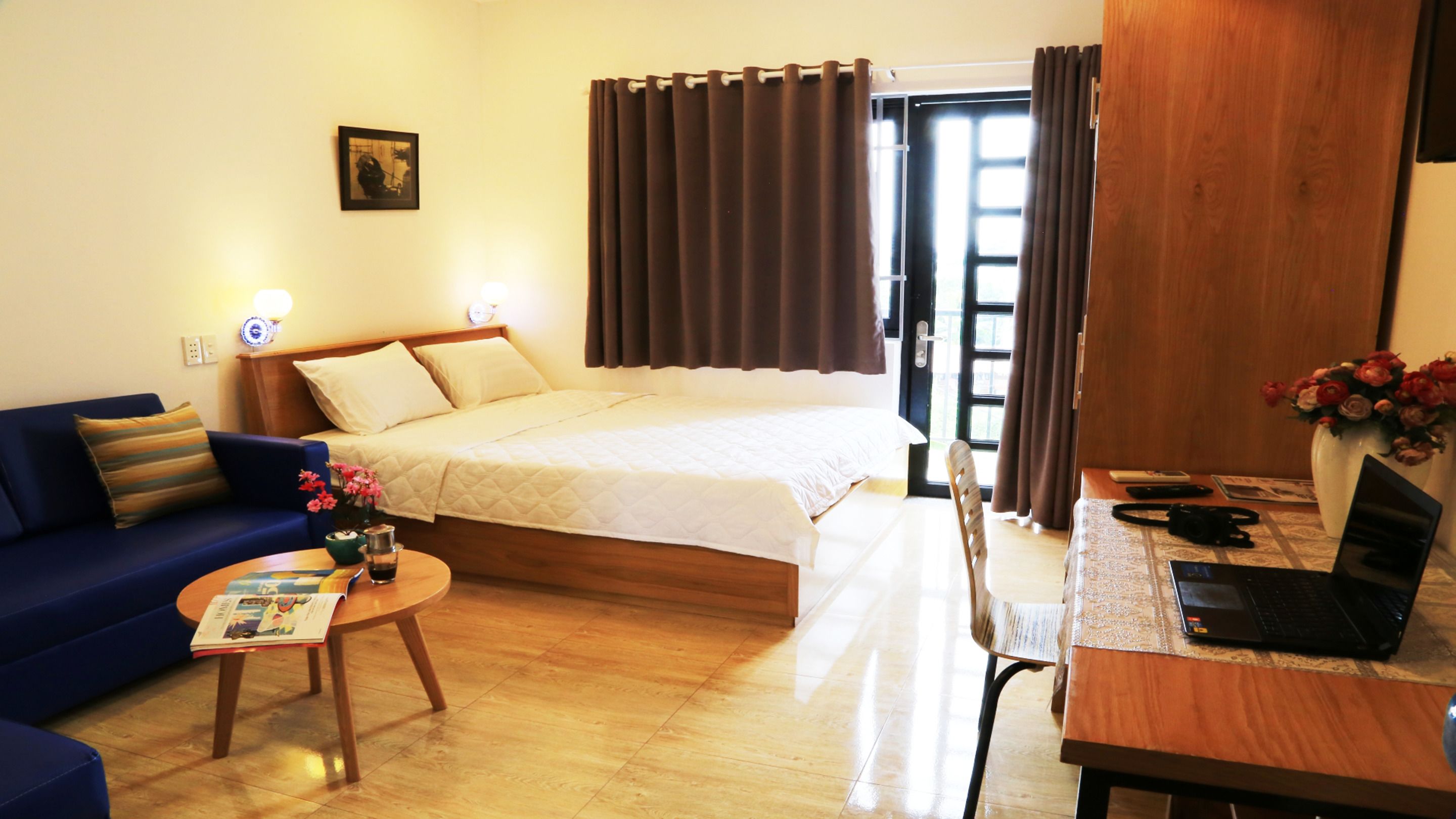 A hotel room with a bed, a sofa, a desk with a laptop, a small table with a magazine, and a vase with flowers.