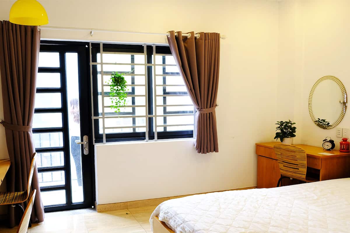 A bedroom with a bed, a desk with a chair, a small table with a plant, a mirror, and a window with curtains.