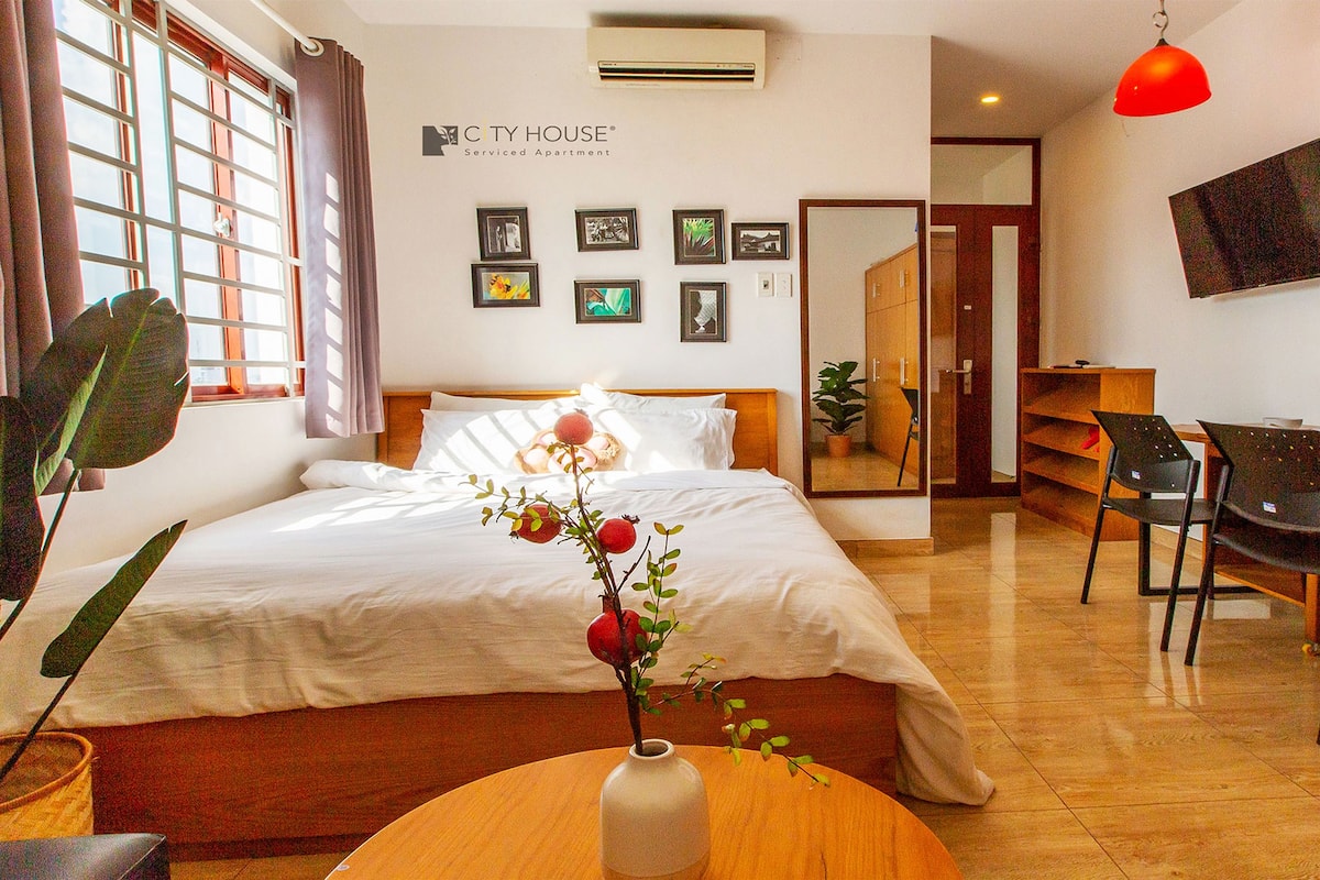A hotel room with a bed, a desk, a chair, a potted plant, and a wall-mounted TV.