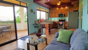 A modern kitchen with wooden cabinets, a microwave, and a refrigerator. There is a dining area with a glass table and chairs, and a living room with a sofa and a decorative vase.