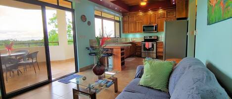 A modern kitchen with wooden cabinets, a microwave, and a refrigerator. There is a dining area with a glass table and chairs, and a living room with a sofa and a decorative vase.