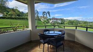 A balcony with a glass table and chairs, overlooking a lush green landscape and a large house.