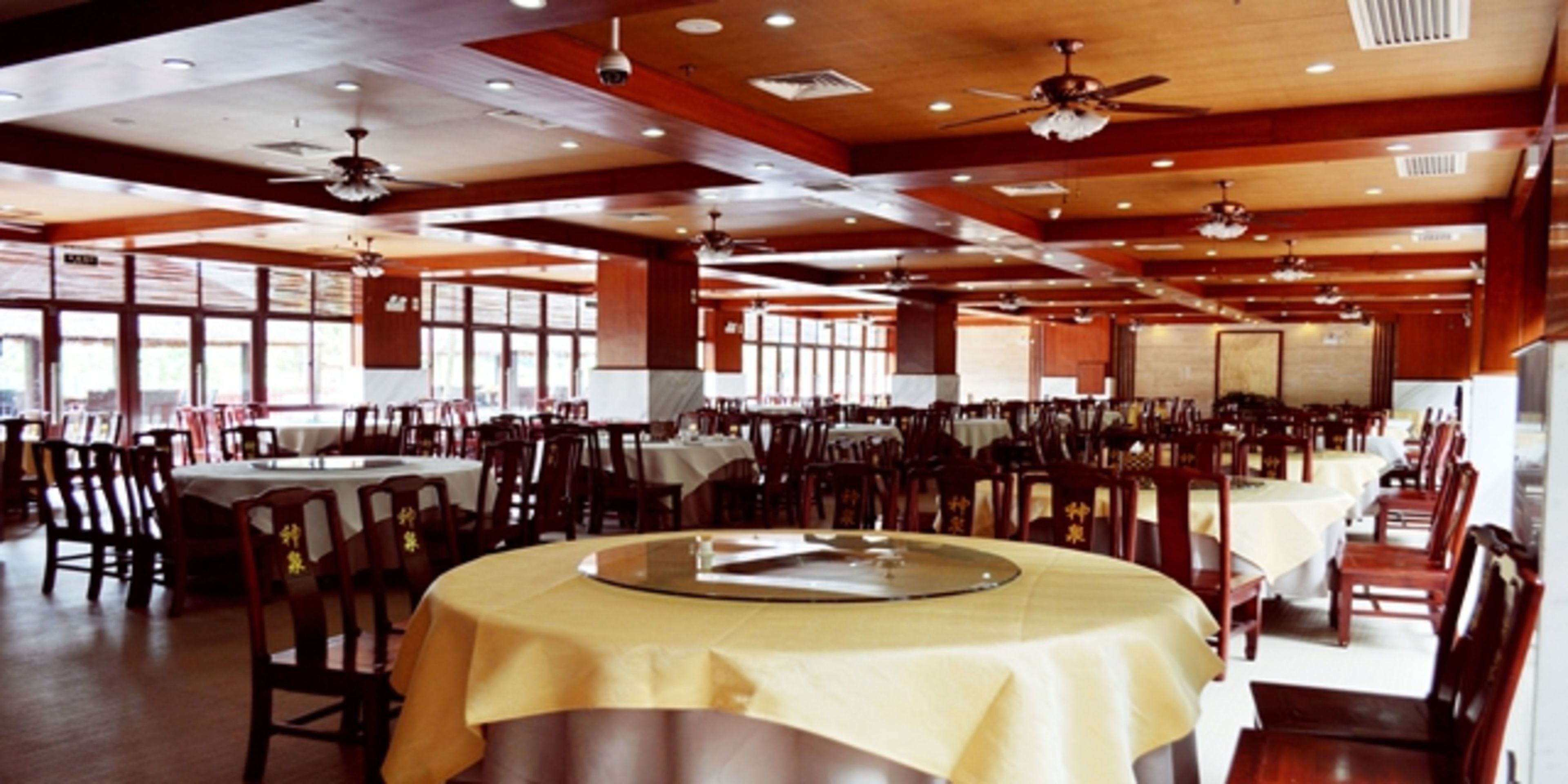 A spacious dining area with multiple tables and chairs, ceiling fans, and large windows allowing natural light.