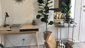A modern living room with a wooden desk, a large potted plant, a leather sofa with patterned cushions, and a glass coffee table.