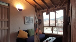 Een rustieke kamer met houten plafond, een bank met kussens, een klein tafeltje met spullen, een raam met uitzicht op gebouwen en een wandlamp.
