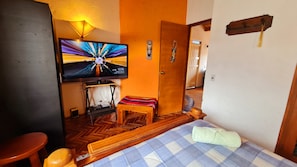 A hotel room with a television on the wall, a wooden bed, a small wooden table, and a bench.
