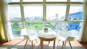 A room with a large window, two white chairs, a small round table with a glass top, and a view of a cityscape and greenery outside. - Indigo Homestay (Magong)