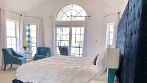 Un dormitorio con una cama grande, dos sillas, una mesita con un florero, y vistas a través de un ventanal.
