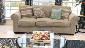 Un salón con sofá beige, cojines estampados, una mesa de centro de vidrio y vistas a una piscina y edificios a través de ventanas amplias.