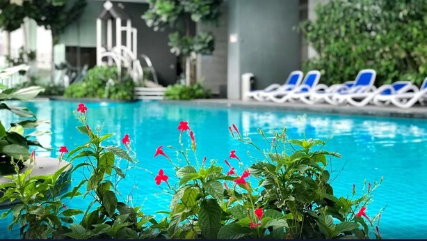 Une piscine intérieure à l’eau bleue, entourée de plantes vertes et de fleurs rouges. Des chaises longues sont disposées le long du bord de la piscine.