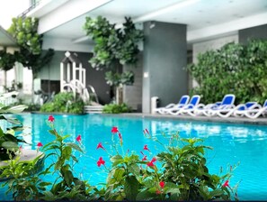 En indendørs swimmingpool med blåt vand, omgivet af grønne planter og røde blomster. Der er liggestole langs poolkanten.
