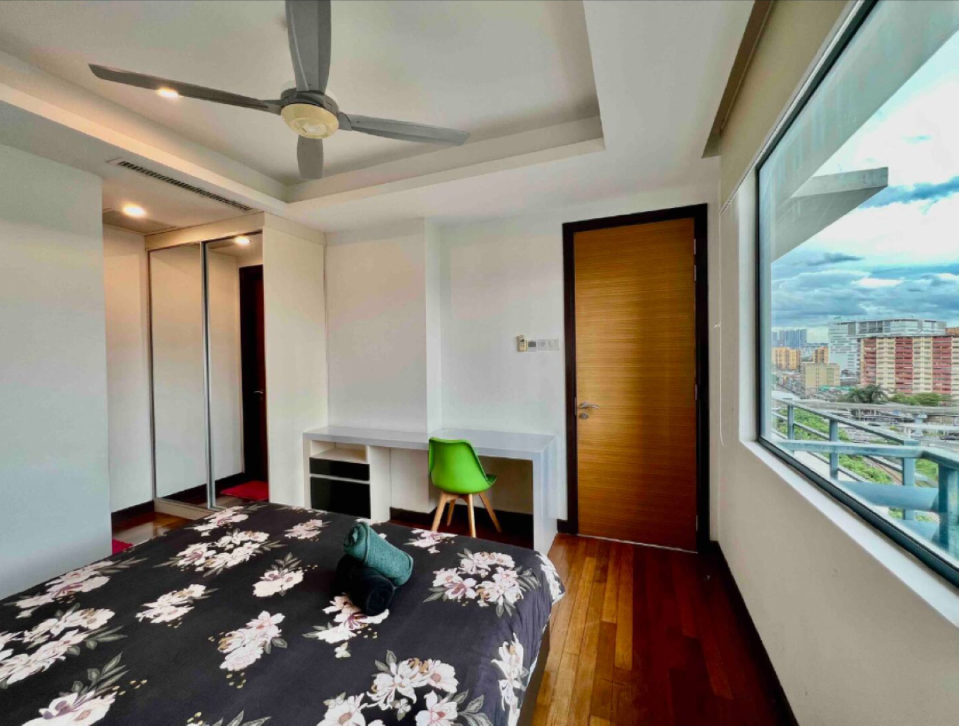 Un dormitorio moderno con una cama, una silla verde, un ventilador de techo y un ventanal con vistas a la ciudad.