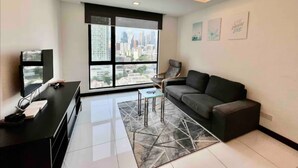 Un salon moderne avec une grande fenêtre, un canapé noir, une table basse et un téléviseur posé sur un meuble.