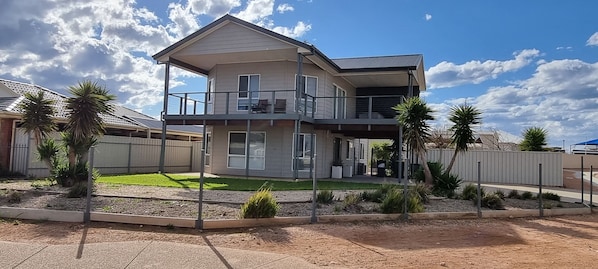 A two-story house with a balcony, surrounded by a landscaped garden with palm trees.
