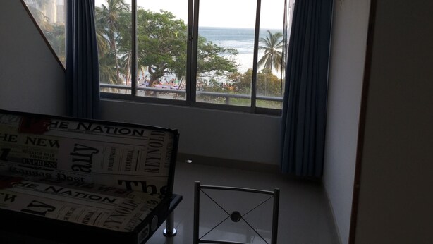 Una habitación con un gran ventanal que da a una playa, una silla de metal y una pila de periódicos.