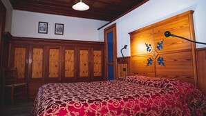 Une chambre avec une armoire en bois, un lit recouvert d’une couvre-lit à motifs et un plafond avec des poutres apparentes.