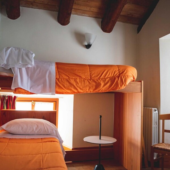 Ein Etagenbett mit orangefarbener Matratze, ein kleiner runder Tisch und ein Stuhl in einem Zimmer mit Holzbalken und einem Fenster.