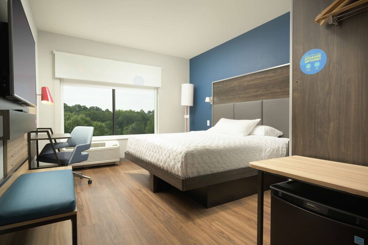 A modern hotel room with a bed, bedside table, desk, chair, and a view of the outdoors.
