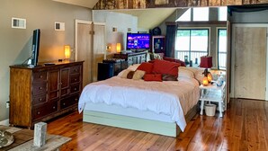 Quarto com teto inclinado, vigas de madeira, uma cama grande, televisão e cômoda.