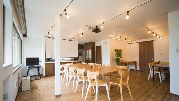 A spacious living room with a large wooden dining table, multiple chairs, and a television. - Hostel KIKO (Sendai)