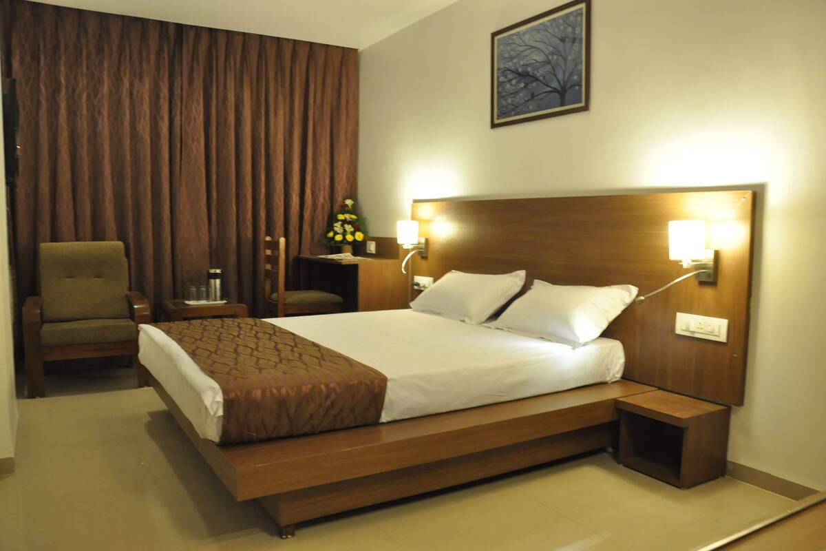 Habitación de hotel con una cama grande, cabecera de madera, dos mesitas de noche, una silla y una cortina.