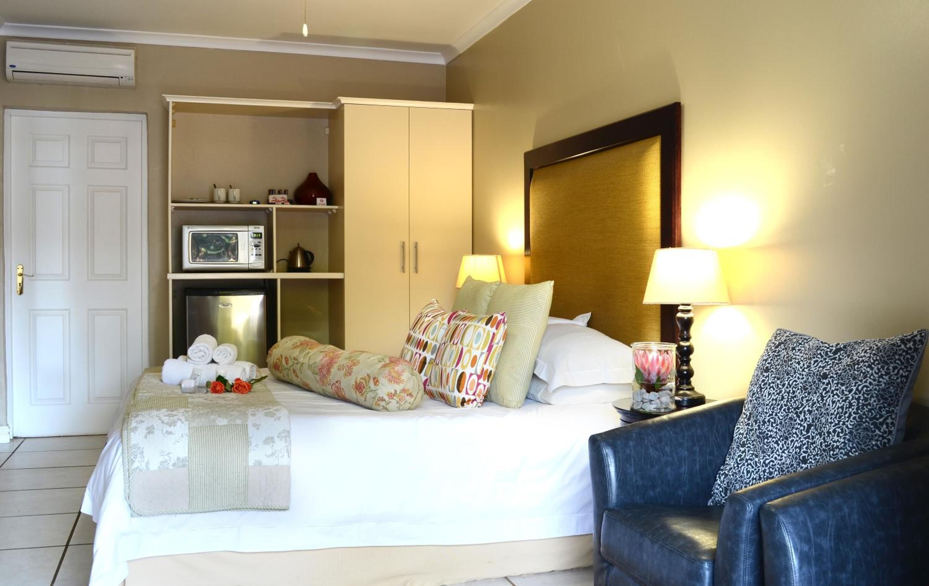 Habitación de hotel con cama, armario, microondas, refrigerador, cafetera y mesita de noche con lámpara y vela.