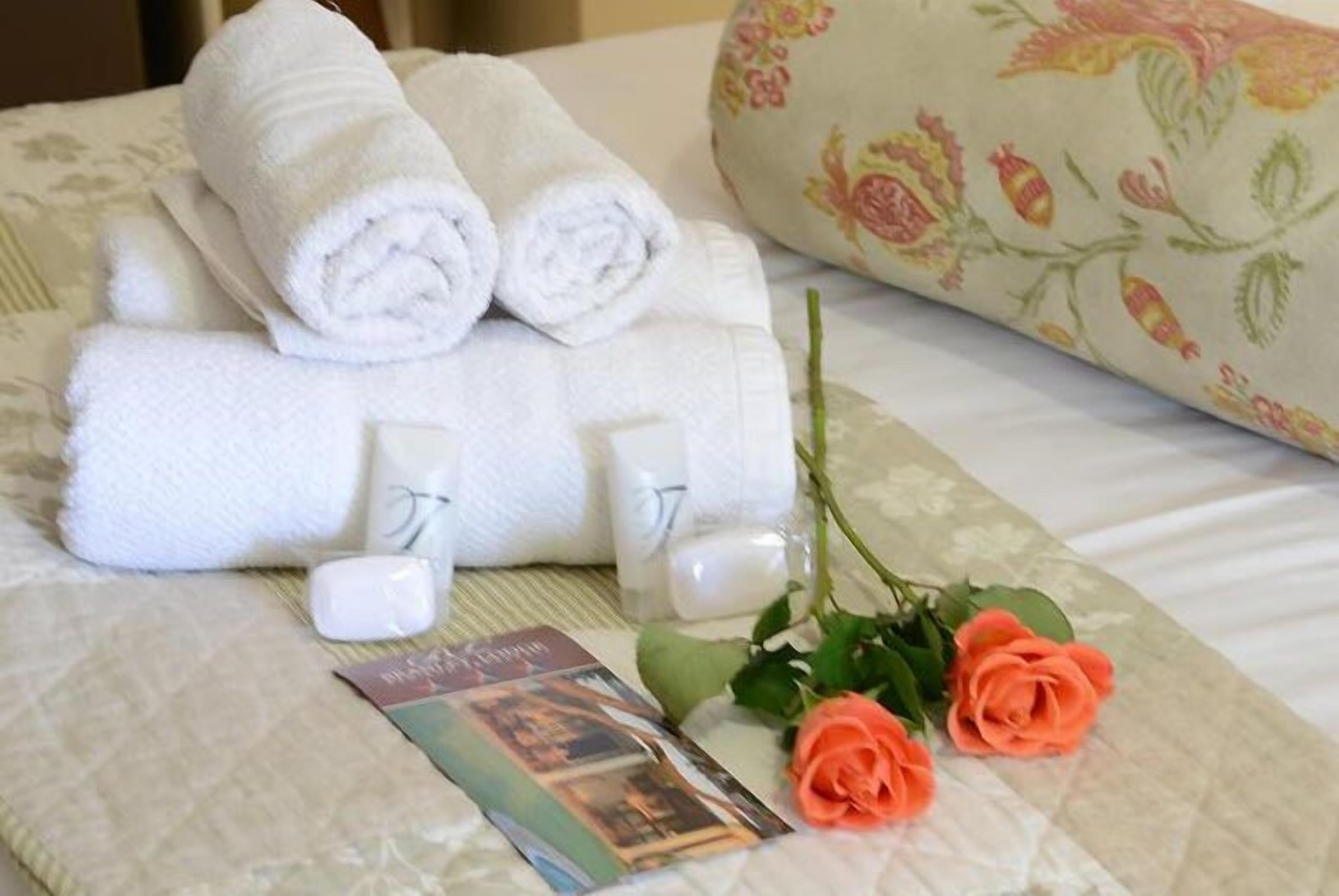 Un lit bien fait, avec des serviettes blanches pliées en rouleau, un coussin à motifs floraux, une rose et un livre.