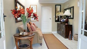 A hallway with a patterned sofa, a small round table with a vase of flowers, a mirror, and a cabinet with lamps. - Branley Lodge (Hillcrest)