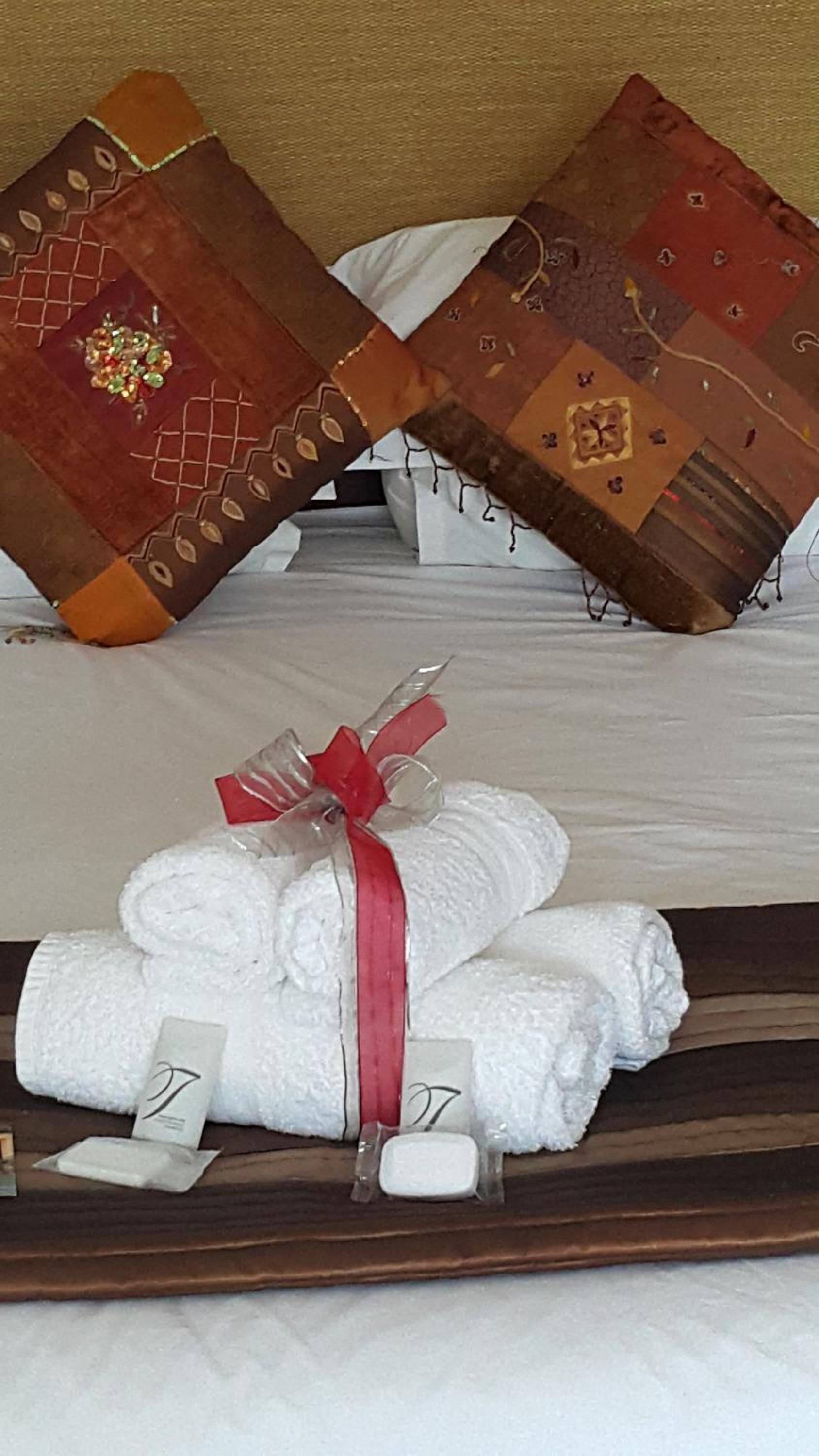 Una cama bien hecha con sábanas blancas y un lazo rojo, acompañada de cojines decorativos y una bandeja con toallas dobladas y artículos de aseo.