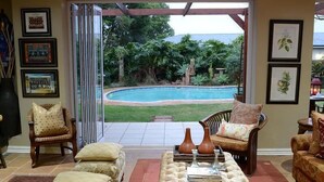 A living room with a view of a swimming pool, a wooden chair, a cushioned sofa, and a coffee table. - Branley Lodge (Hillcrest)