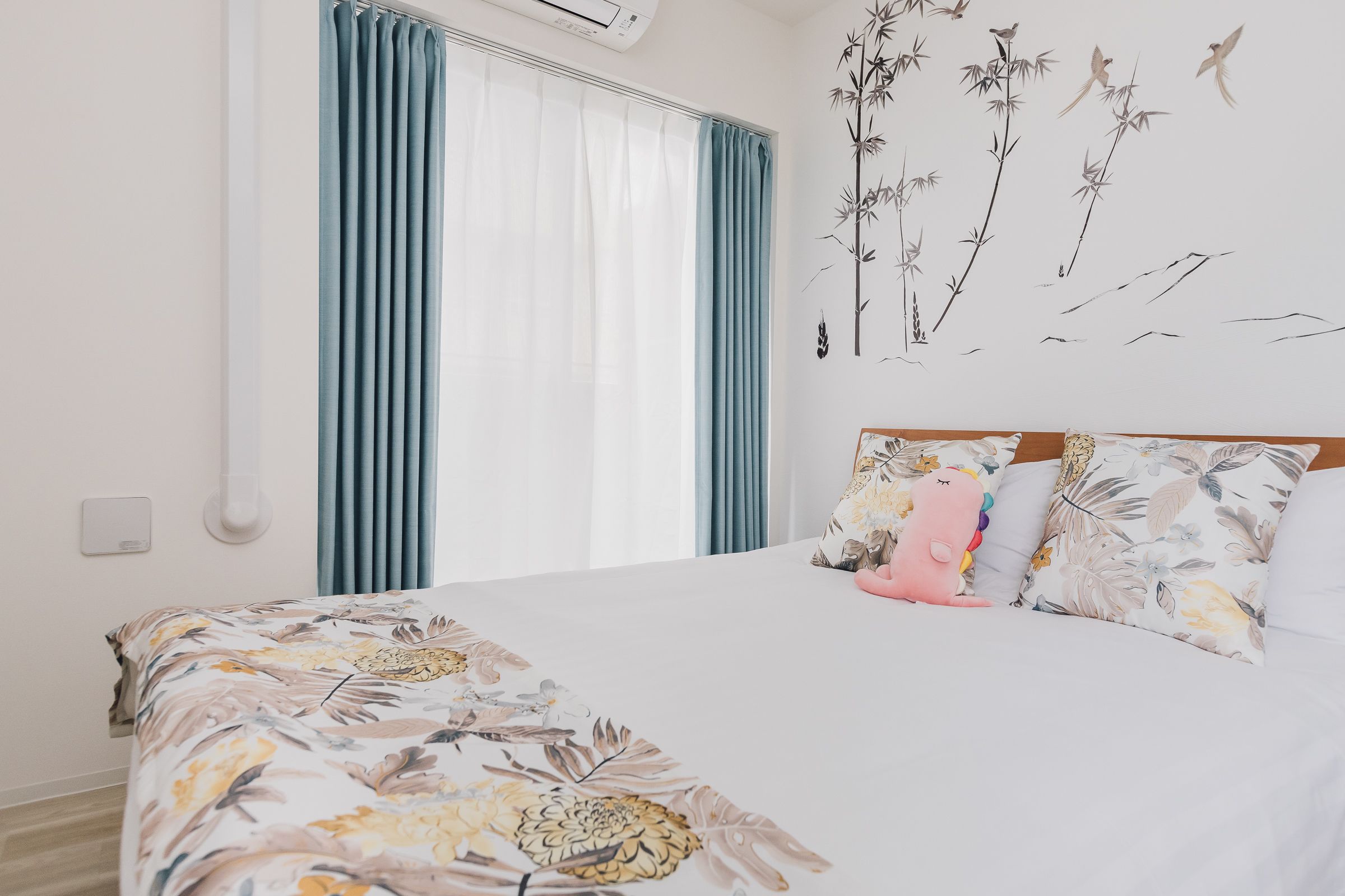 A bedroom with a large bed, floral bedding, a wall-mounted air conditioner, and a window with curtains.