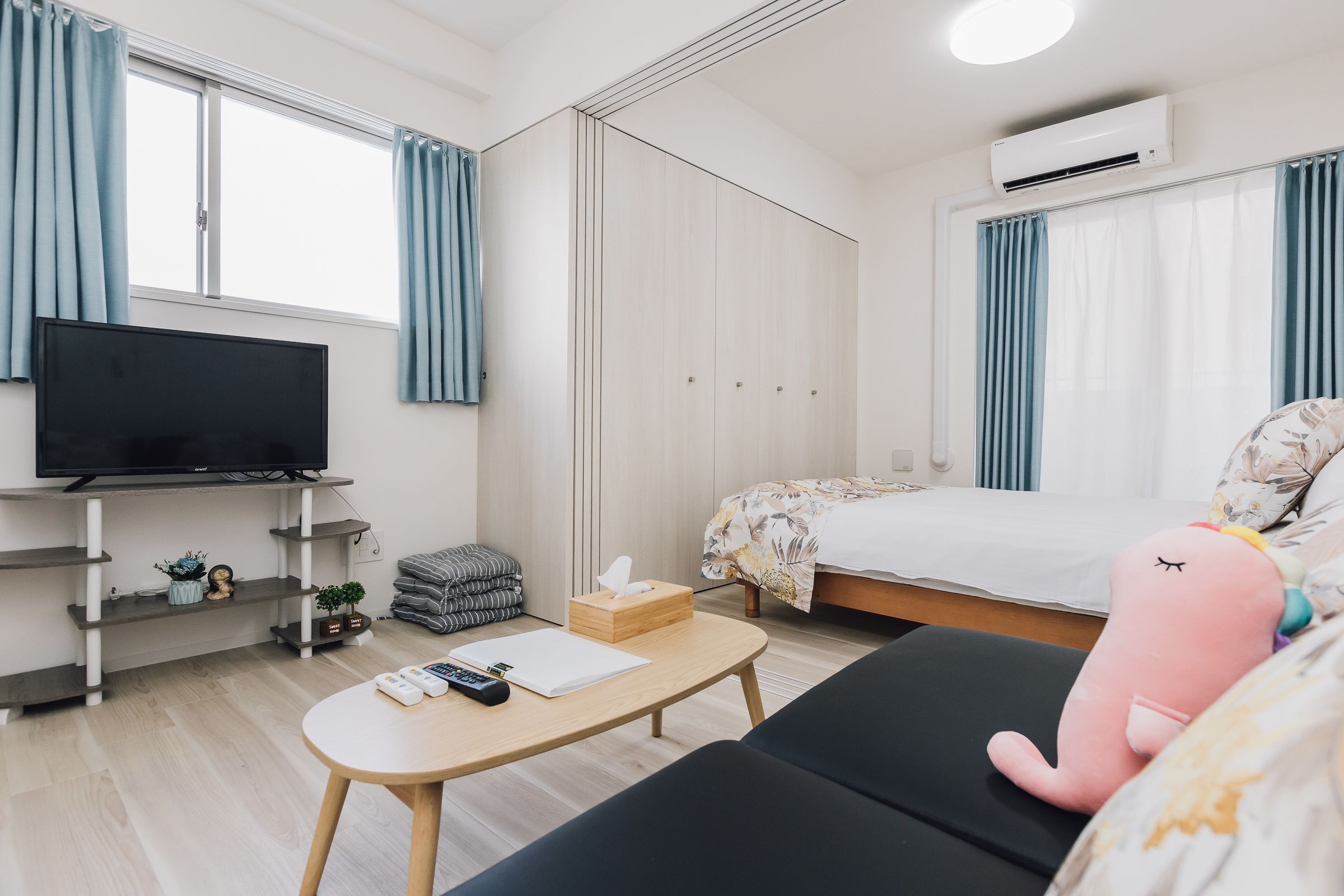 A modern hotel room with a television, a bed with a floral bedspread, a small table with a remote control and a notebook, and a pink stuffed toy on the bed.