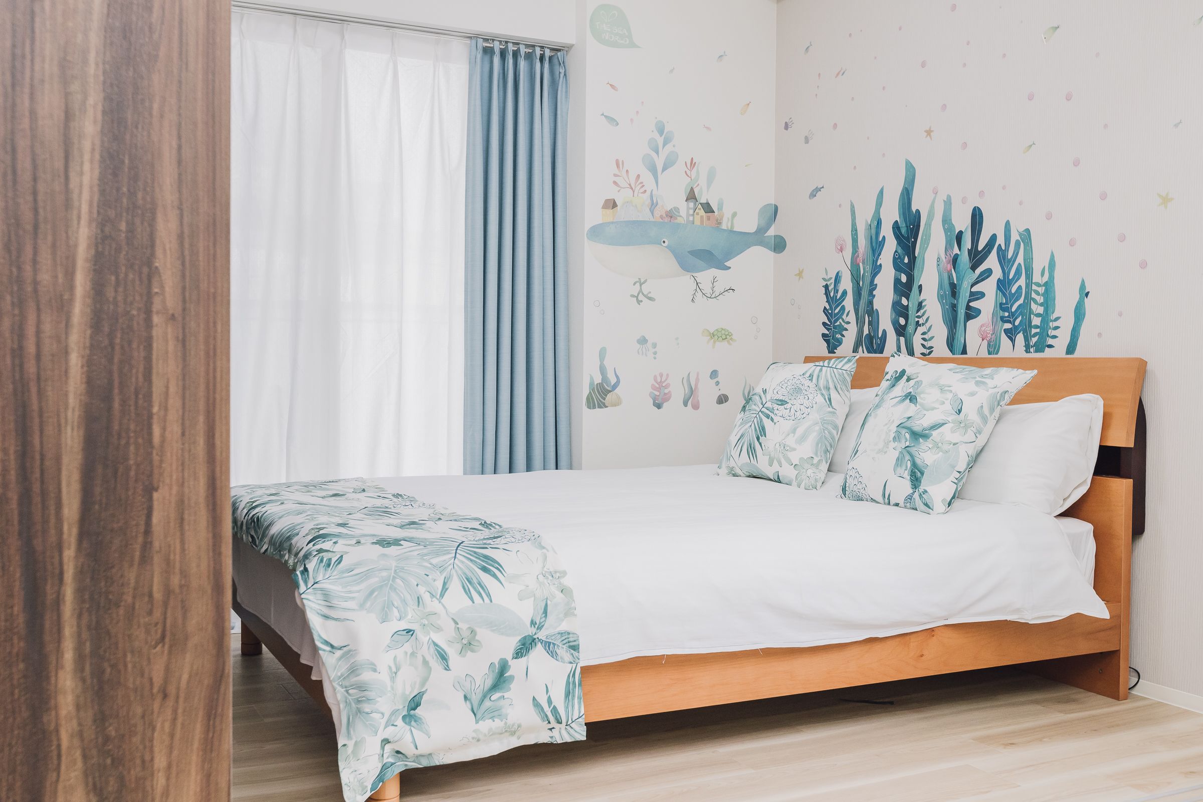 A bedroom with a wooden bed, white bedding, a tropical leaf-patterned throw, and a wall mural of underwater scenery.
