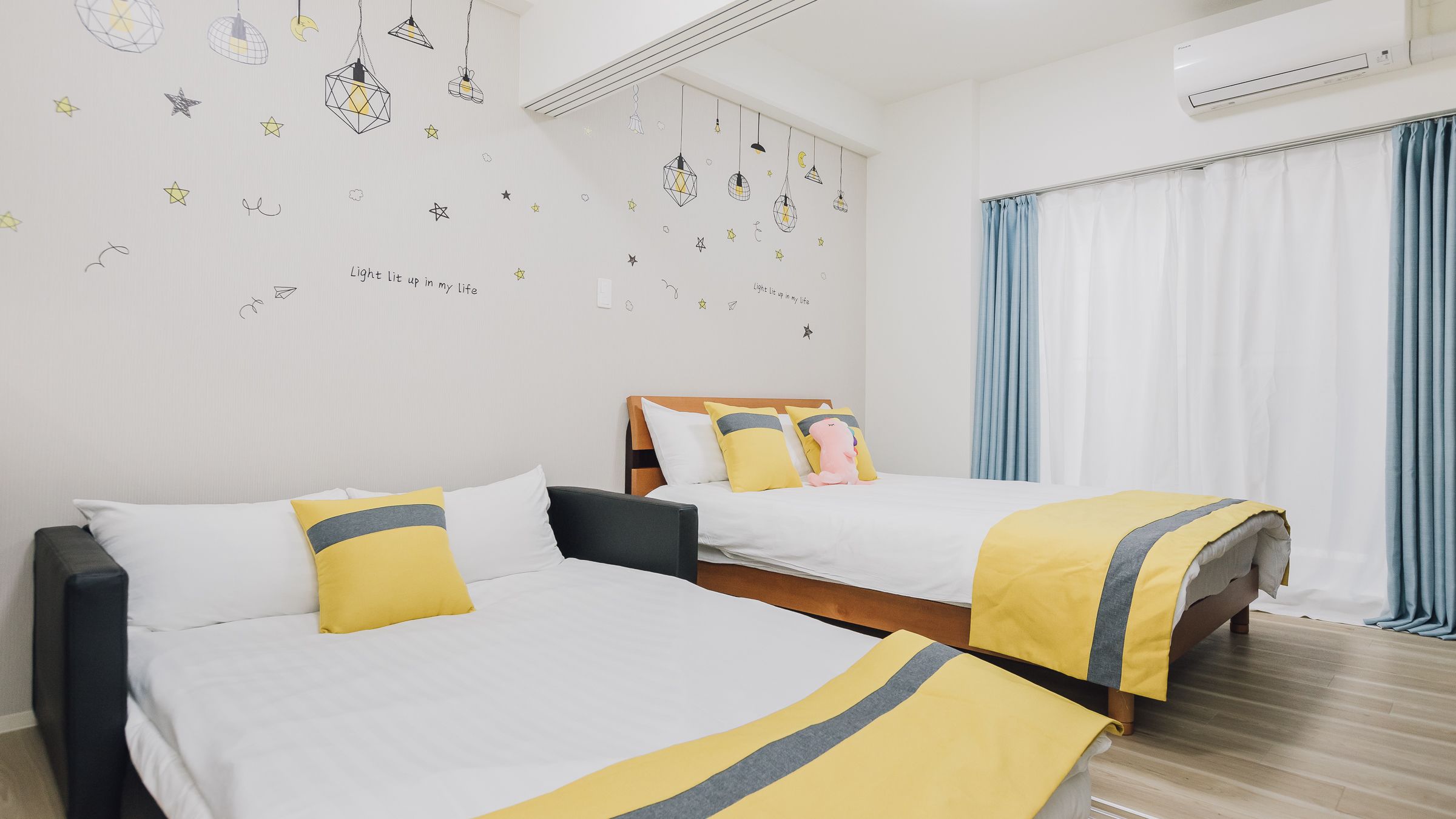 A hotel room with two beds, each with white bedding and yellow accents, a wooden headboard, and a wall decorated with stars and geometric shapes.