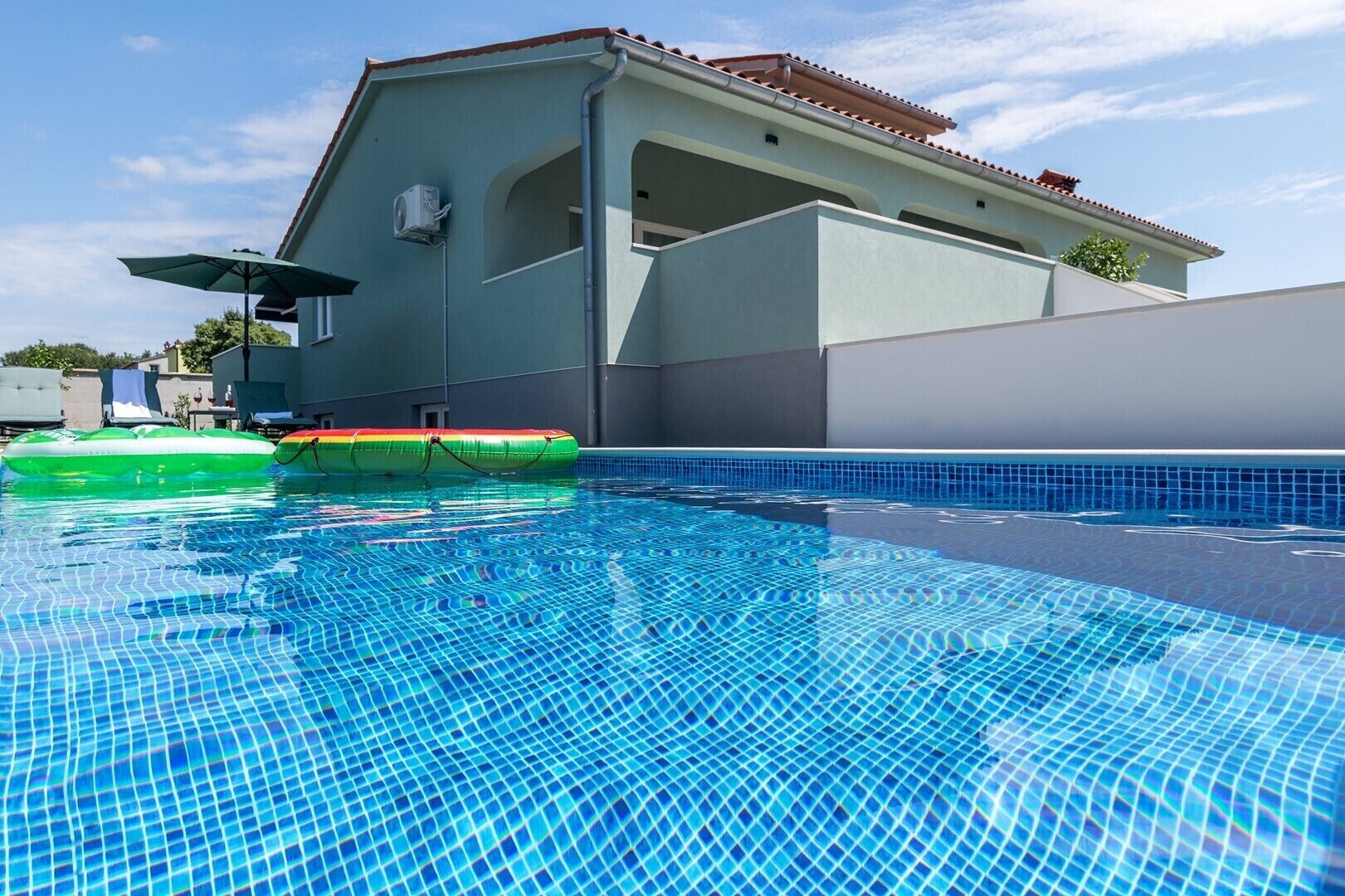Une piscine aux carreaux bleu clair, un flotteur gonflable vert et, au fond, un bâtiment avec un toit recouvert de tuiles.