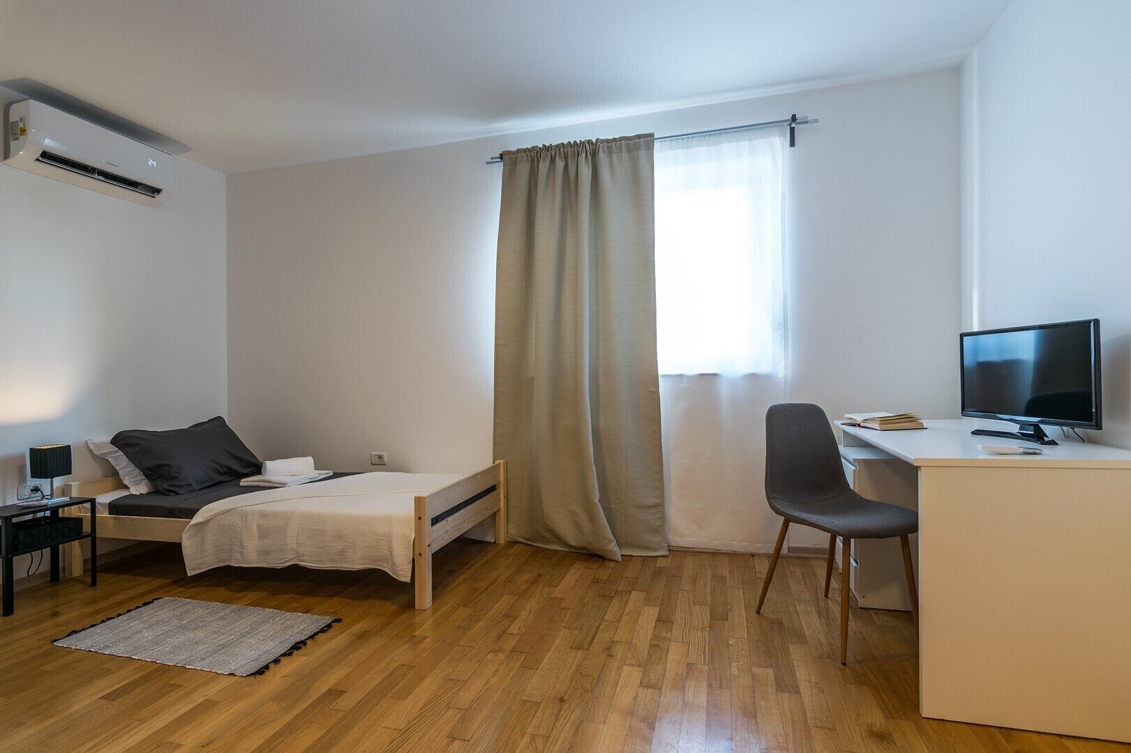 Een kleine, opgeruimde kamer met een bed, een bureau met een computer, een stoel en een raam met gordijnen.