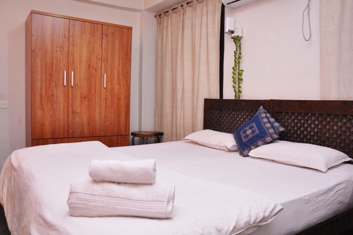 A neatly made bed with white linens and folded towels, a wooden wardrobe, a small round table, and a potted plant.