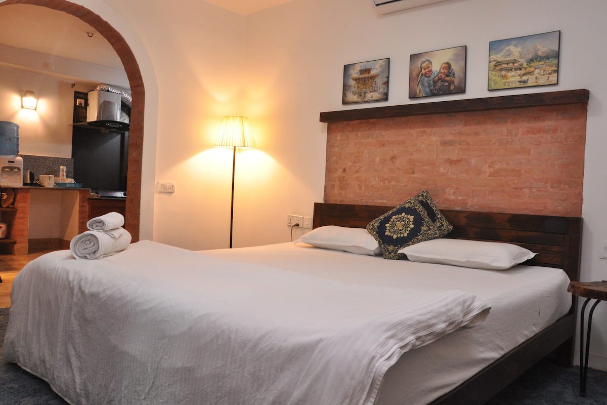 A hotel room with a bed, a lamp, a TV, and a kitchen area visible through an archway.