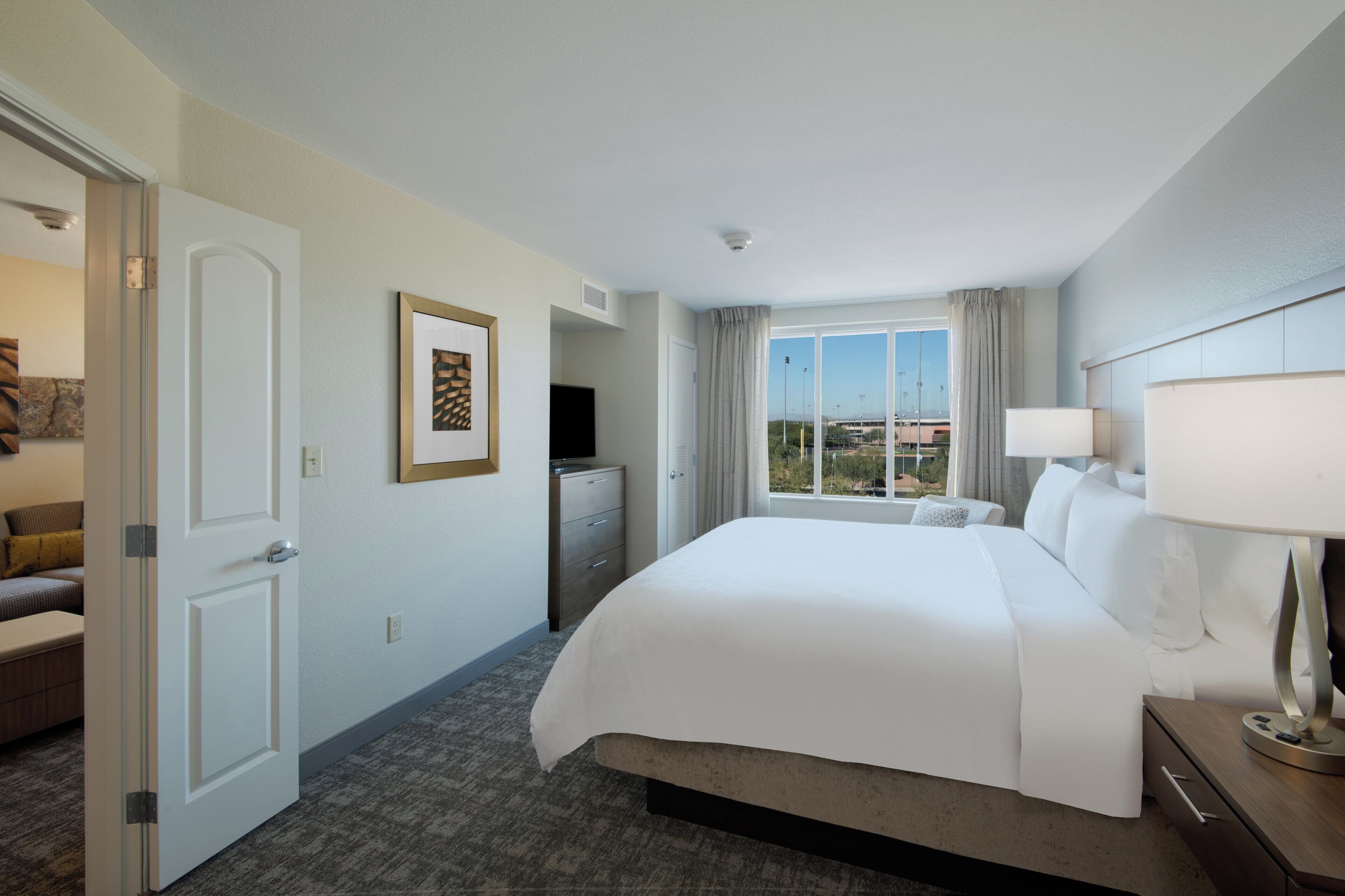 Quarto de hotel com uma cama grande, uma escrivaninha com luminária, uma televisão e uma janela com cortinas.