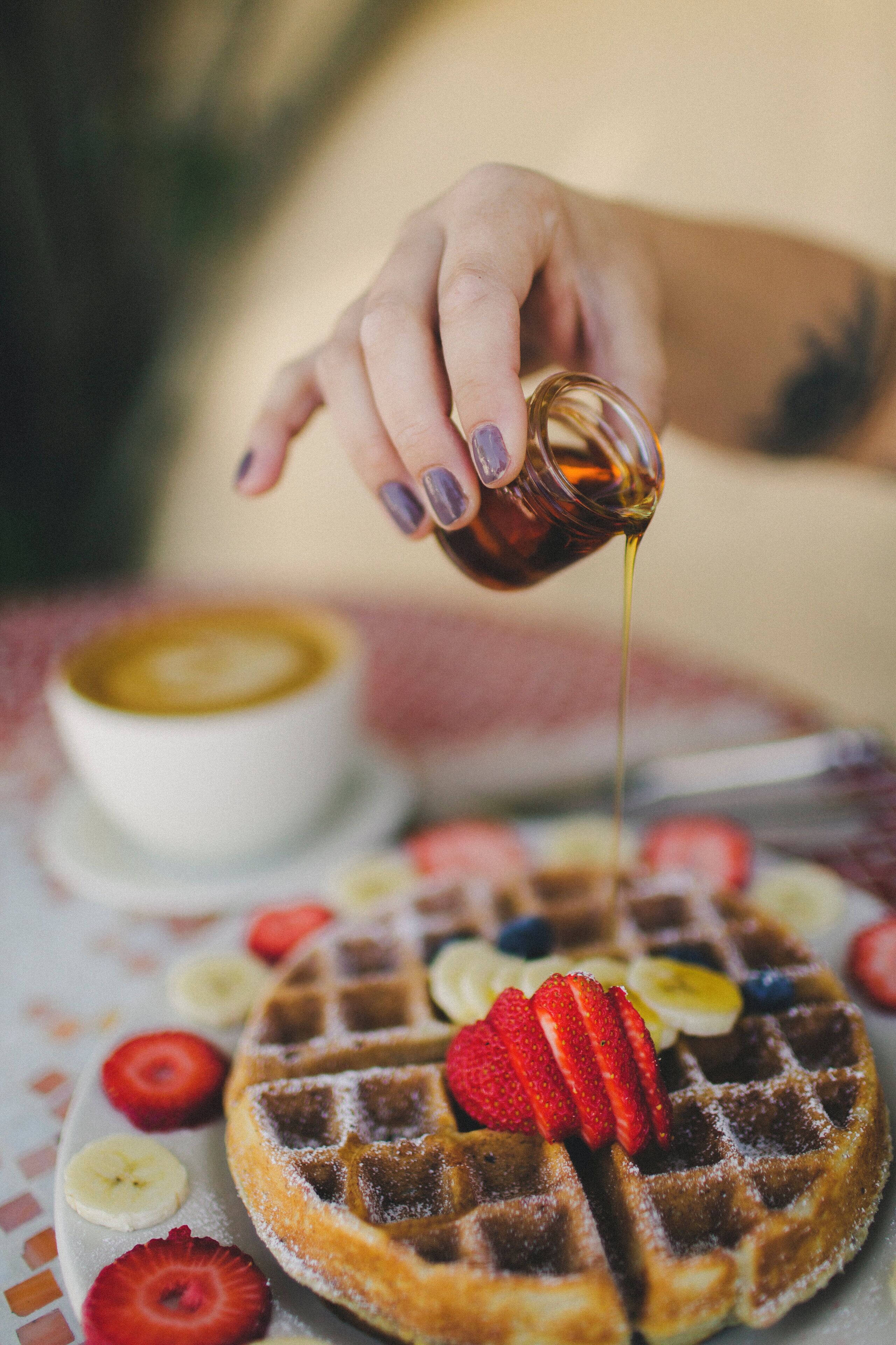 Una mano sirviendo jarabe sobre un waffle con fresas y una rodaja de plátano.